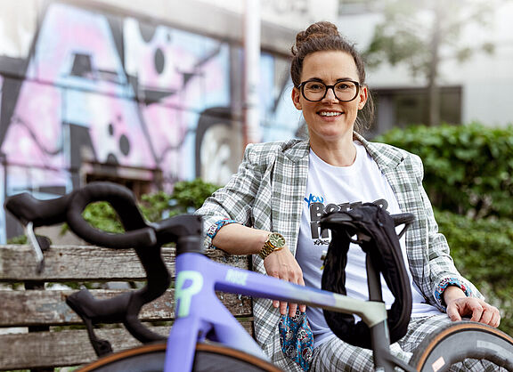 Eine Frau sitzt entspannt auf einer Holzbank neben einem violetten Rennrad vor einer Graffiti-Wand