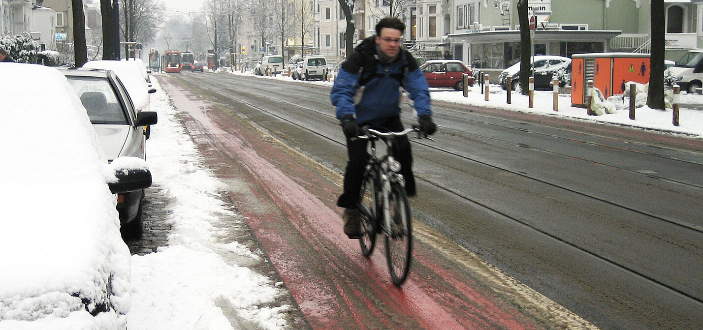 Ein Radfahrer in Winterkleidung fährt auf einem rot markierten Radfahrstreifen neben einer Straße, der weitgehend von Schnee befreit ist, während am Straßenrand und auf geparkten Autos Schnee liegt.
